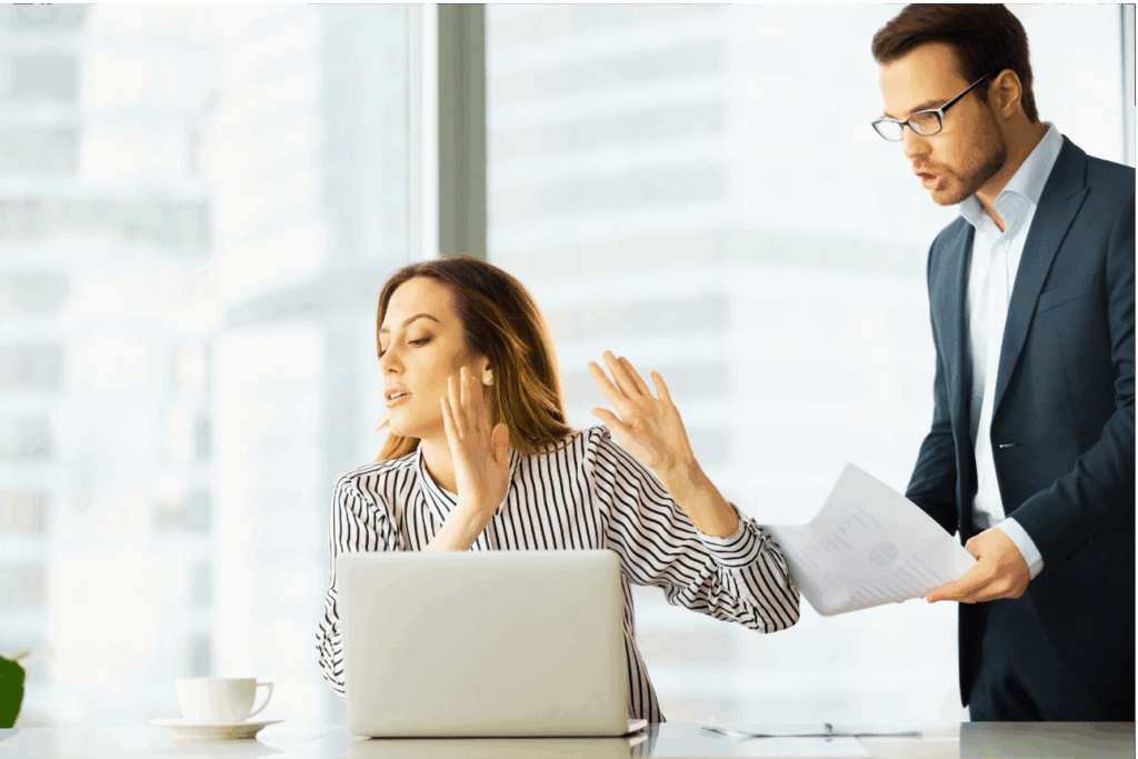 Zwei Personen in einem modernen Büro. Eine Person sitzt an einem Schreibtisch mit einem Laptop und einer Tasse und hebt eine Hand, als ob sie einen Widerstand signalisiert. Die andere Person steht neben dem Schreibtisch, hält einige Dokumente und schaut die sitzende Person an. 