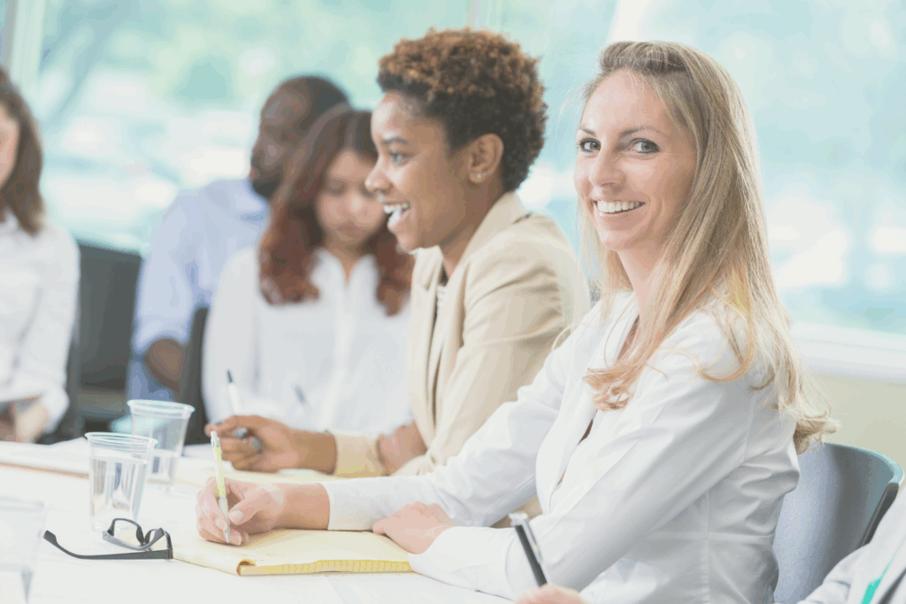 Das Bild zeigt Frauen, die bei einer Weiterbildung teilnehmen. Mehrere Frauen sitzen am Tisch. Die Frau im Vordergrund hat einen Block vor sich und einen Stift in der Hand. Sie lächelt in die Kamera. 